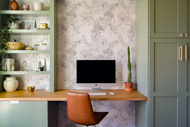 A modern office space. Olive-painted built in shelves and cabinets come together with a natural wood desk. The wall is covered in a cream colored wallpaper with an outlined floral pattern.