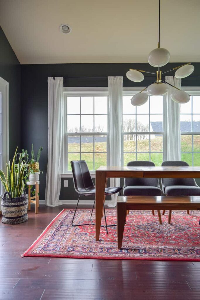 A newly renovated dining room features dark colored walls and large windows with white linen curtains. The wood dining room table is surrounded by three chairs and a wood bench, under a modern globe chandelier.