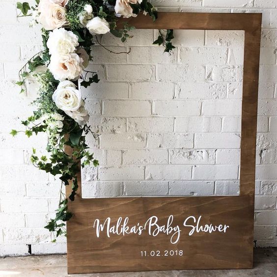 An oversized wooden polaroid frame stained a dark brown color. White and green floral garland is wrapped around one corner. On the bottom of the frame, white text reads "Malika's Baby Shower, 11/02/2018"