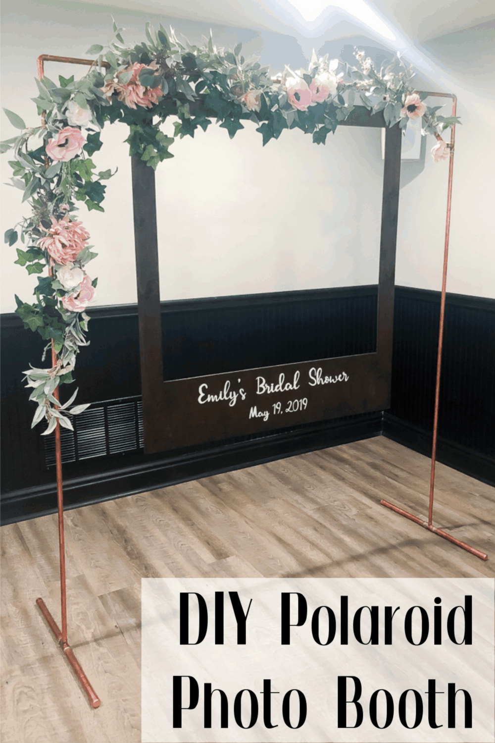 A creative DIY polaroid photo booth made with a copper pipe arch decorated with flower garland. A wooden polaroid frame hangs from the arch, and says "Emily's Bridal Shower, May 19, 2019" in white paint.