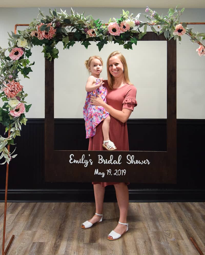 A young blond woman wearing a pink dress, holding a toddler girl wearing a pink floral dress, stands behind a wood polaroid picture frame attached to a copper arch decorated with floral garland.
