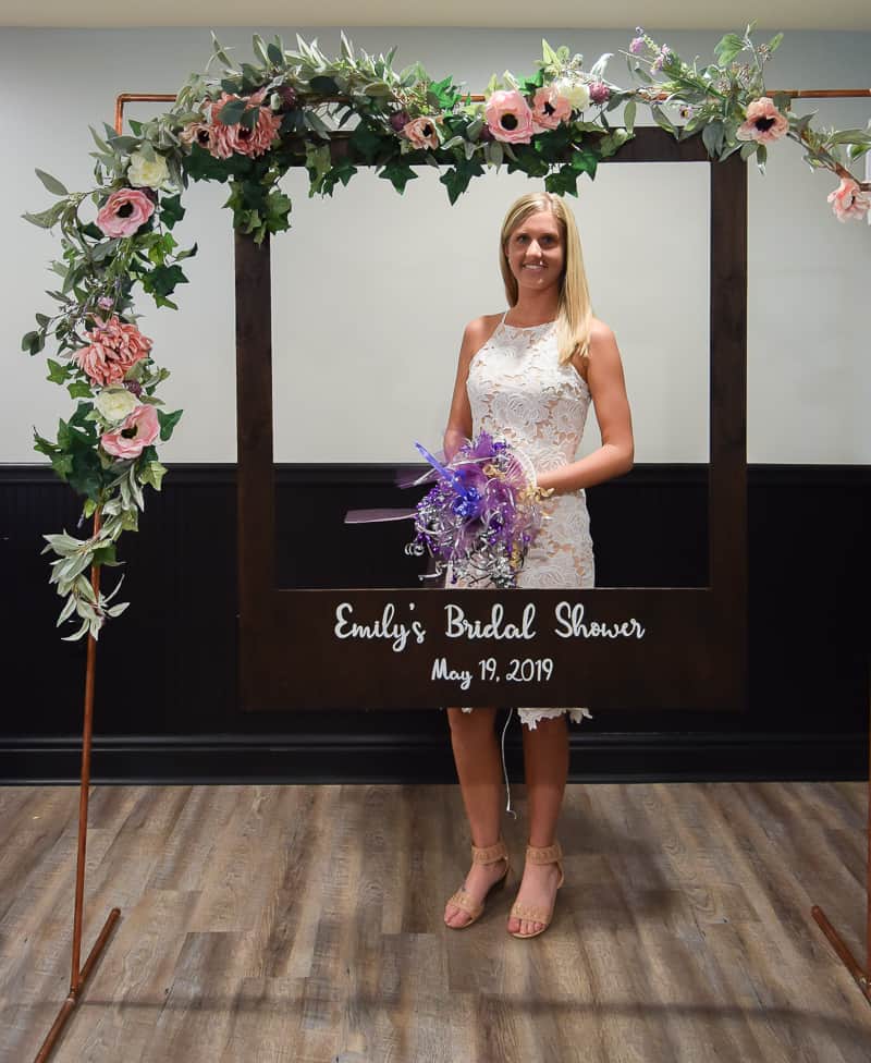 A young blond woman wearing a white dress is holding a ribbon bouquet. She stands behind a DIY polaroid photo booth made with a copper arch and pink and white flower garland.