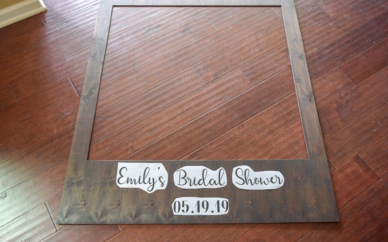 A dark wood polaroid photo booth frame lays on a hardwood floor. Text printed on paper sits across the bottom of the frame and reads "Emily's Bridal Shower, 05.19.19"