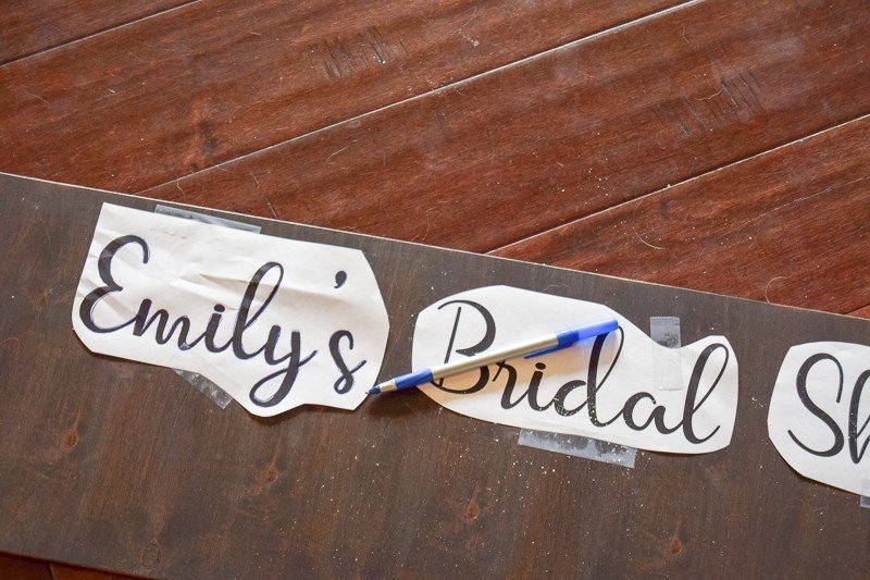 A blue ink pens sits across pieces of paper taped to a wood frame. The papers read "Emily's Bridal Shower"