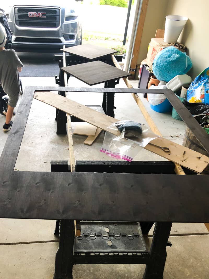 The process of staining the large polaroid photobooth frame with a dark brown wood stain. the frame rests on a workshop work horse set up.