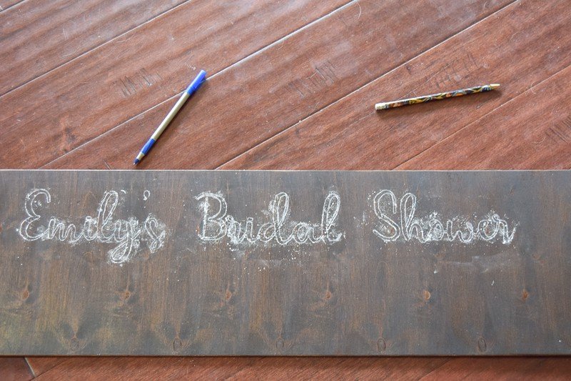 The words "Emily's Bridal Shower" are traced onto the bottom of a dark wood polaroid picture frame using chalk. Just the outline of the letters is visible.