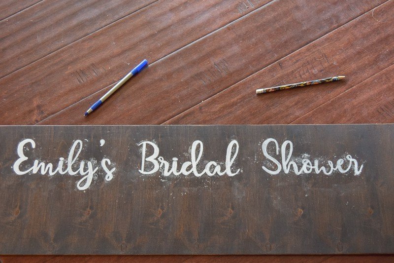 The words "Emily's Bridal Shower" are filled in with white paint, painted onto the bottom of a dark wood polaroid picture frame.