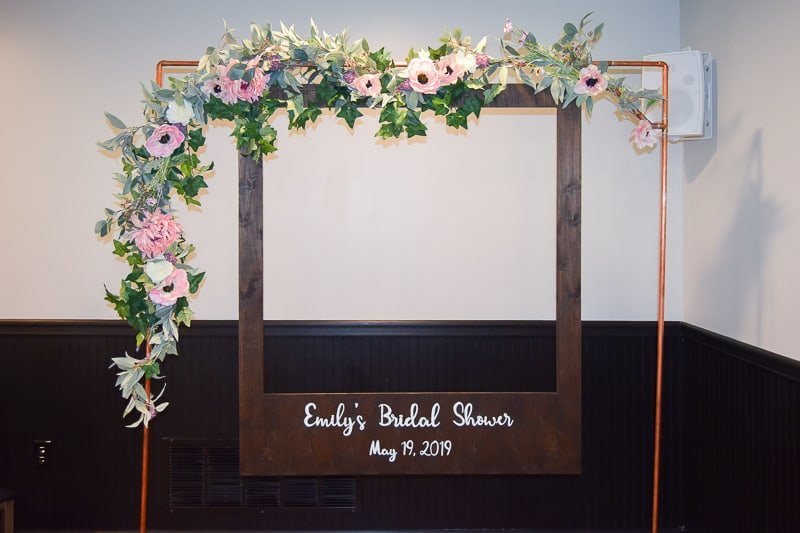 A large copper pipe arch decorated with a strand of garland with pink and white flowers and green leaves wrapped around the top. An oversized polaroid picture frame hangs from the copper pipe arch that says "Emily's Bridal Shower"