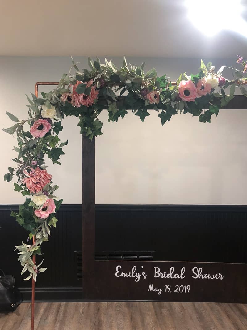 A closer look at the flower garland wrapped around the copper arch. The garland has white and pink flowers with light and dark green leaves, surrounding a dark wood stained polaroid picture frame.
