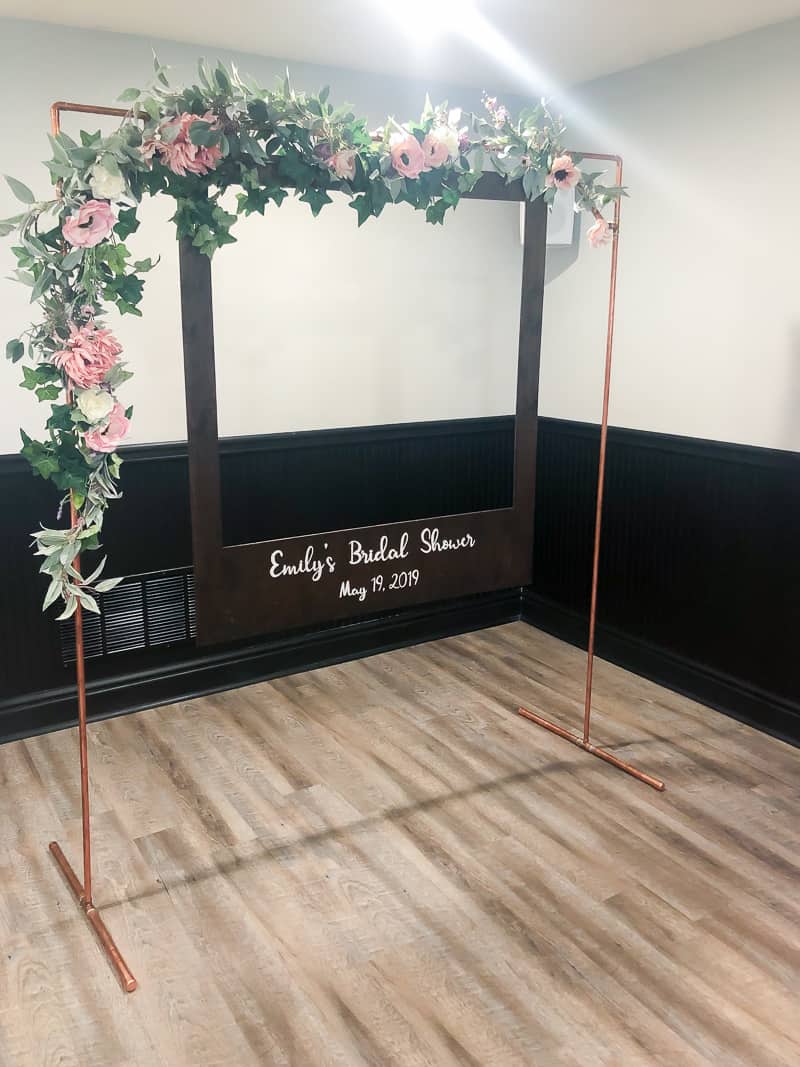 Another angle of the copper arch photo booth with flower garland and an oversized polaroid picture frame hanging in the center.