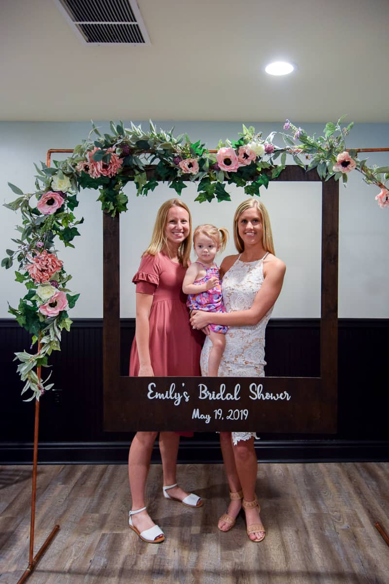 Two young blond women stand together with a little girl in their arms, behind the oversized polaroid picture frame hanging from a DIY copper arch, decorated with pink and white flower garland.