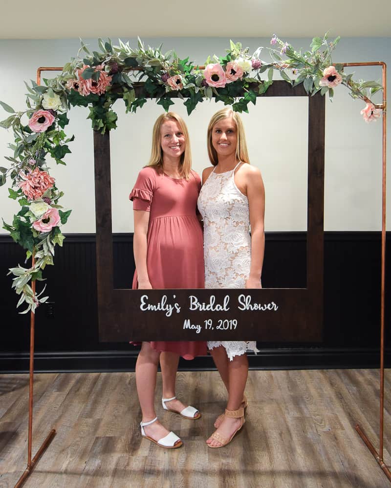 Two blond women - one wearing a pink dress and one wearing a white dress - stand behind a DIY polaroid photo booth, decorated with pink and white floral garland.