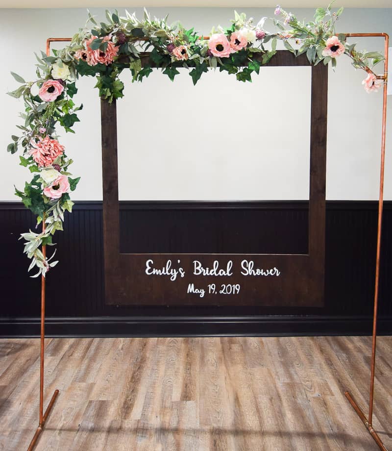 The completed DIY polaroid photo booth, with a wooden polaroid picture frame hanging from a copper arch, decorated with a pink and white floral garland.