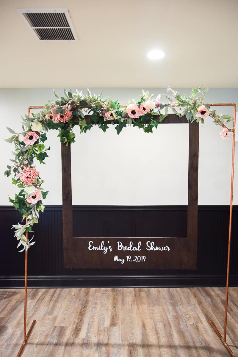 A large polaroid photo booth made from copper pipes, with a pink and white floral garland wrapped around the top and side of the arch. A dark-stained wood polaroid picture frame hangs from the arch, and says "Emily's Bridal Shower" and the date in white paint.