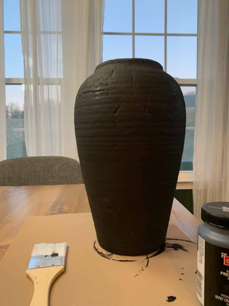 A large vintage vase, painted entirely black, sits on a table while the paint dries.