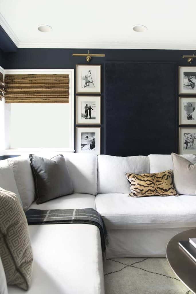 A living room with a white fabric sectional couch and deep navy blue walls, decorated with framed photos. The wall paint color is Naval from Sherwin-Williams.