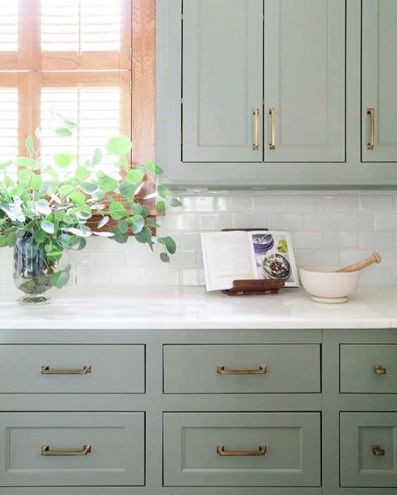 Sage green kitchen cabinets with brass handles, painted Clary Sage, SW 6178 by Sherwin-Williams. The kitchen has white marble countertops and a white subway tile backsplash.