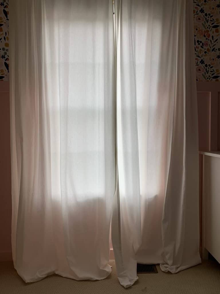 Long floor length white curtains hang over a window, and pool on the carpeted floor. 