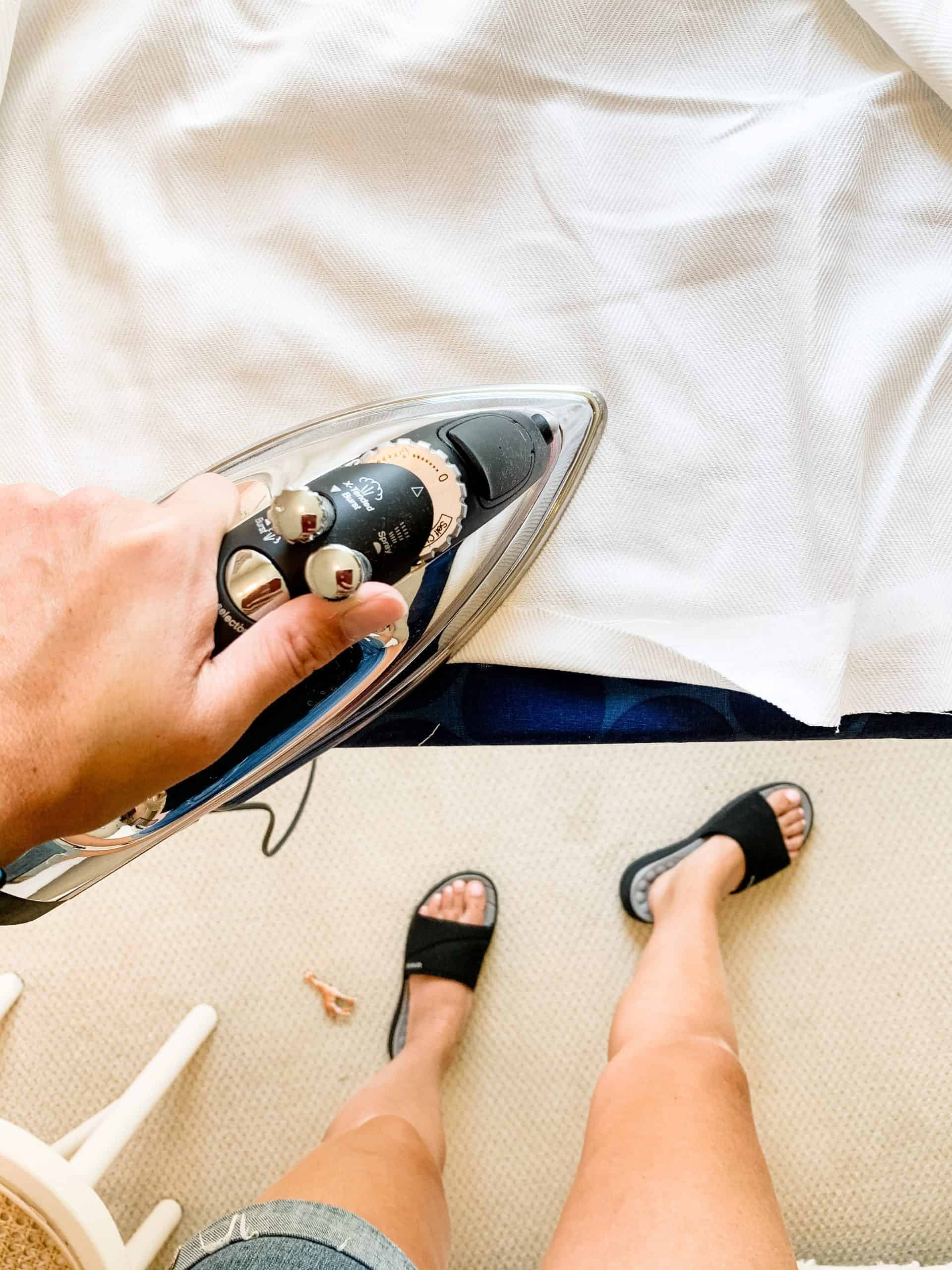 An iron runs across white fabric curtains, creating a no-sew hem at the end of floor-length curtains. A women's legs and feet are visible as her hand holds the iron against the fabric.