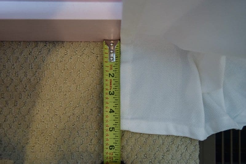 A measuring tape shows five inches of white curtain fabric laying on a carpeted floor.