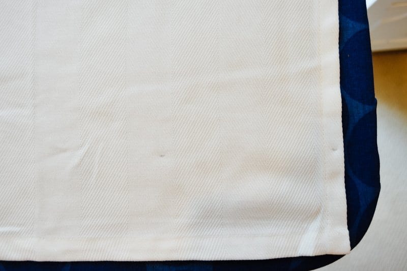 Two barely visible pencil marks on white curtain fabric mark three inches from the curtain hem, laid out flat on an ironing board.