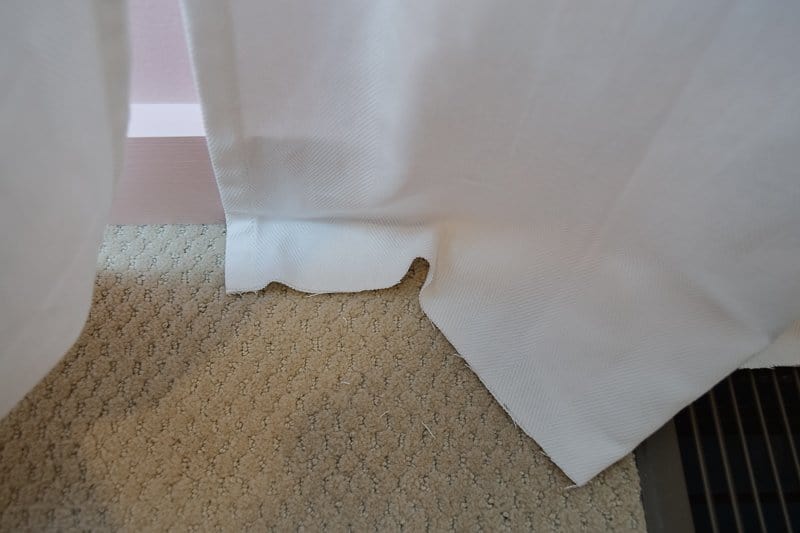 Floor length white curtains hanging in front of a window, with a few extra inches of fabric  falling down on the carpet.
