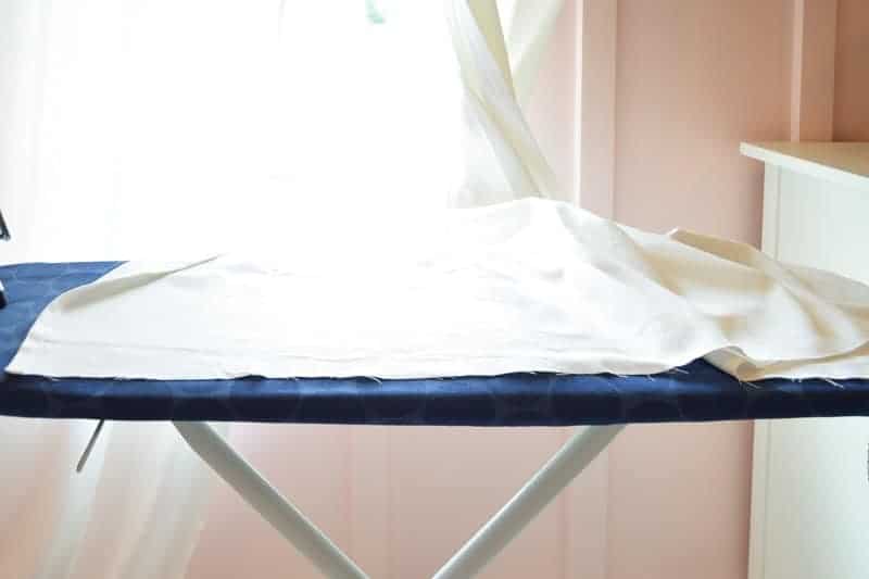 Long white fabric curtains are bunched up onto an ironing board, ready for no-sew hemming.