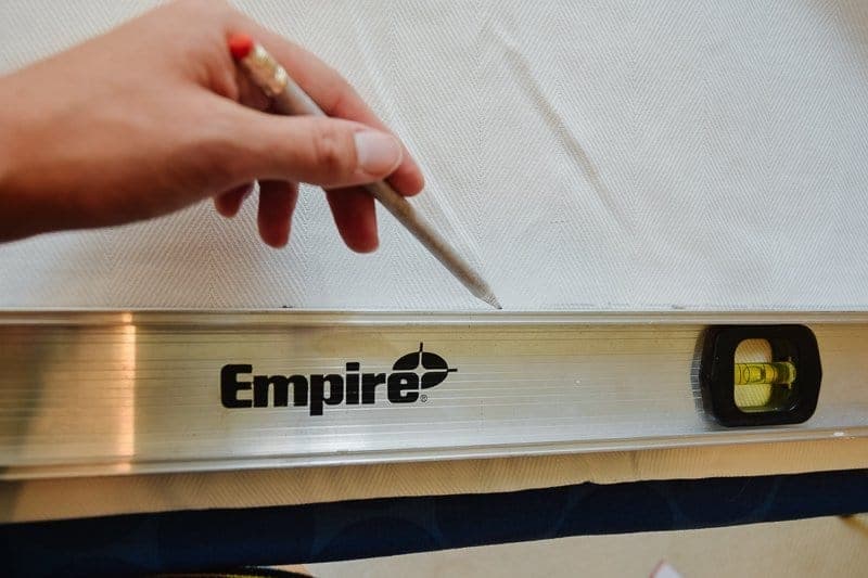 A woman's hand draws a line in pencil along an Empire brand level, creating a single line across the bottom of white fabric curtains.