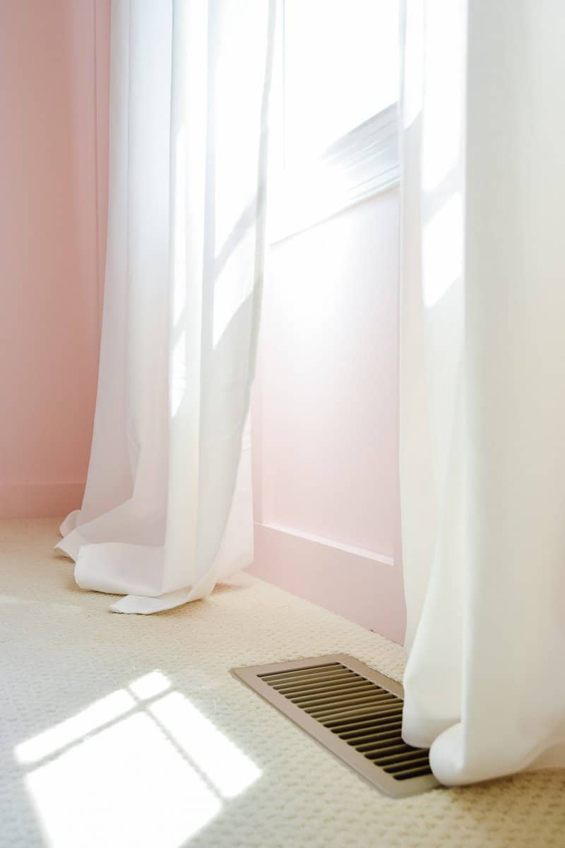 The bottom of two floor-length curtain panels. One white curtain is unhemmed and too long, pooling on the floor. The other panel is hemmed and sits just above the floor.