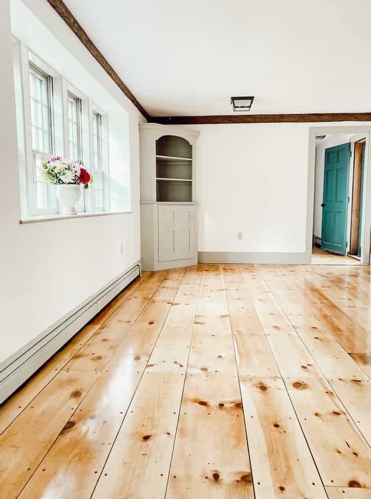 A photo of a white wall painted with Simply White on a large, empty room with cabinet near the window and wooden floor