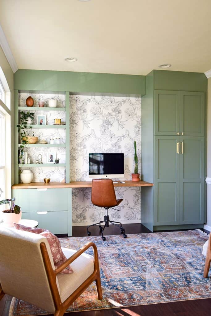 Home office with a green theme using floral line wallpaper in the back of built-in shelves,. cabinetry and desk.