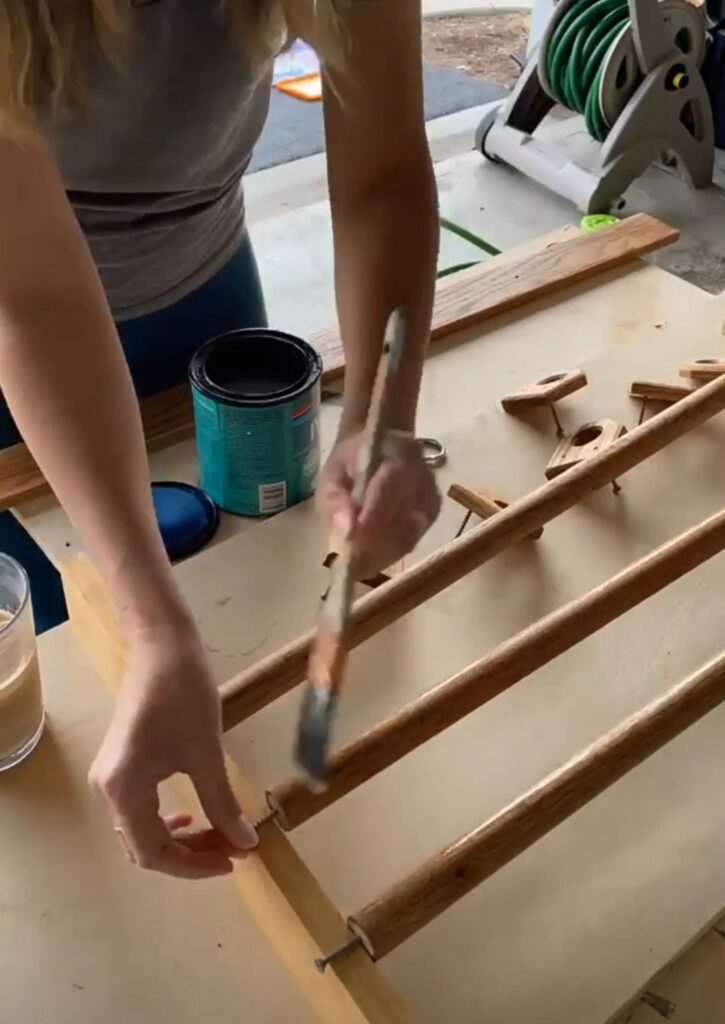 A woman is using a paintbrush to apply polycrylic to a wooden rod.